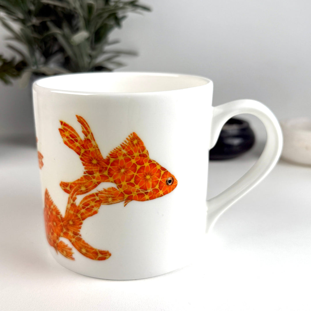 White mug with a goldfish design on a white surface with a plant in the background