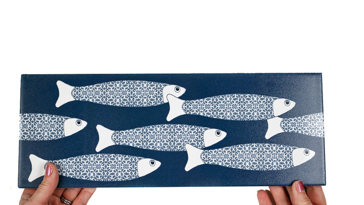A rectangular dark blue decorative tile or tray featuring a pattern of white fish, held by two hands against a plain white background.